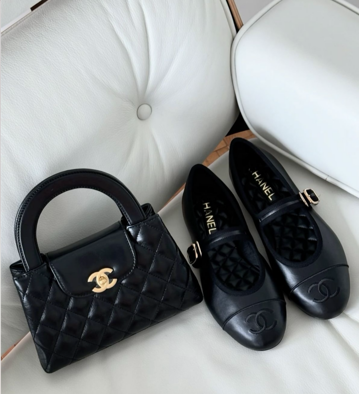 A pair of Chanel Mary Jane flats in Lambskin (FULL SET) from EXCHANGE STORE sits elegantly on a white tufted chair beside a black quilted Chanel bag with gold logo—a must-have display for collectors.