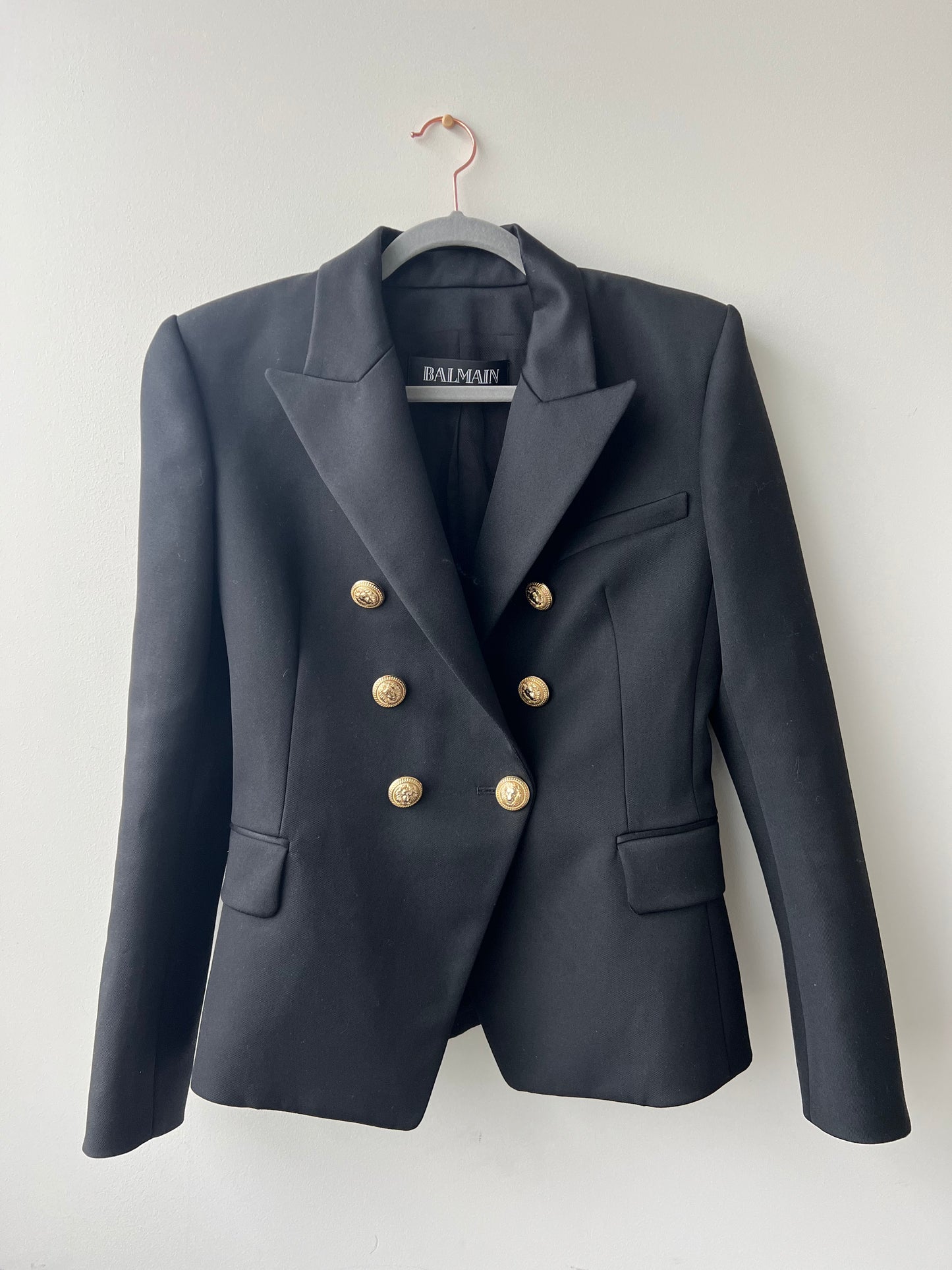A Balmain Blazer Black by EXCHANGE, featuring gold buttons, peaked lapels, and flap pockets, made in Slovakia, hangs on a pink hanger against a white wall.