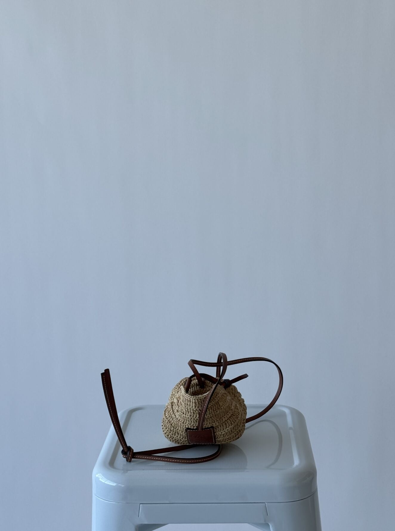 A LOEWE - Cross body shell bag by EXCHANGE, woven in beige with brown leather straps, sits on a light gray stool against a plain background—a pristine collector’s piece with no visible wear.