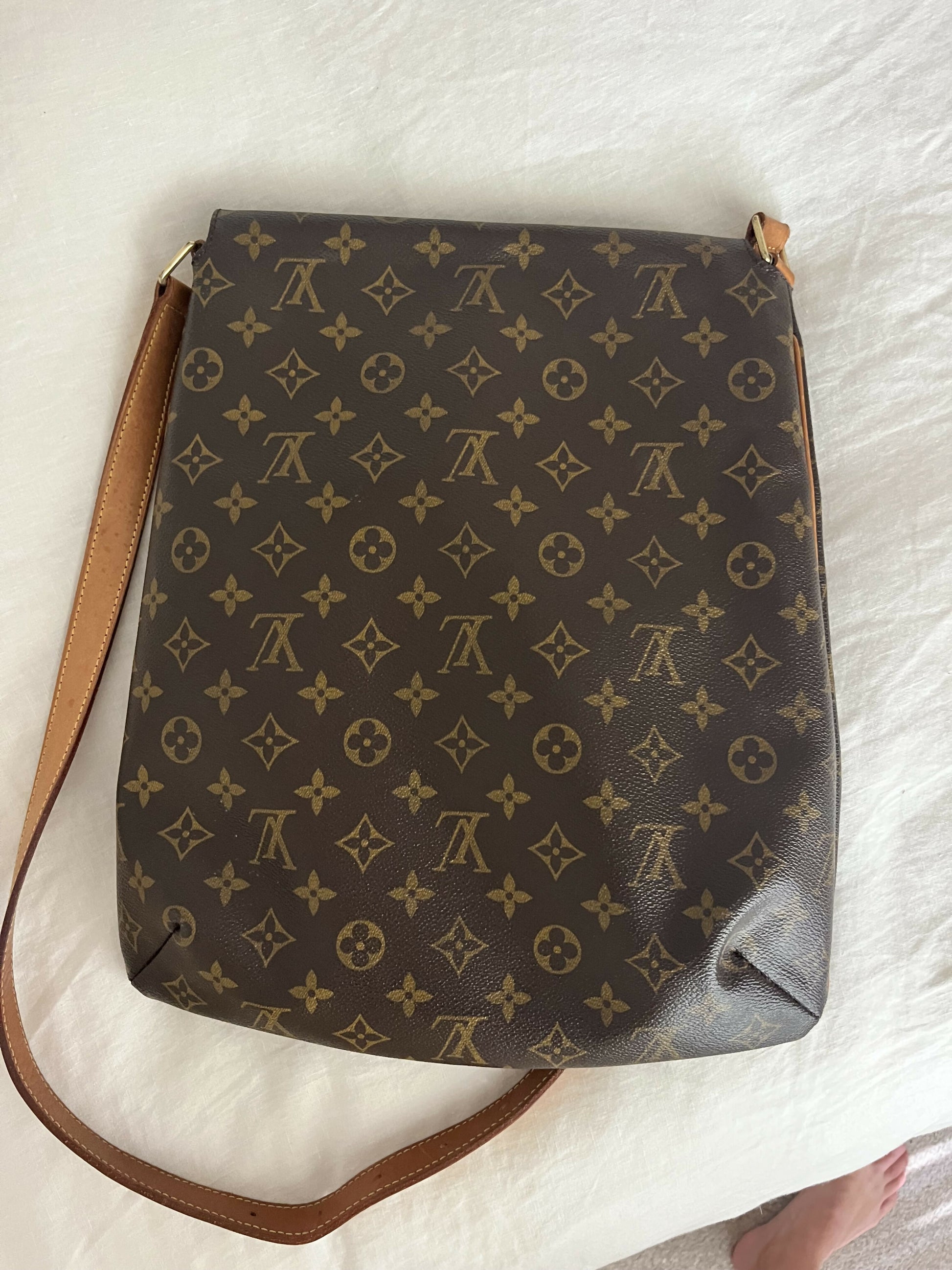 An authenticated Louis Vuitton Monogram Musette shoulder bag by EXCHANGE with a monogram pattern and tan leather strap sits on a white surface, showing wear on the bottom right corner. A bare foot is partially visible in the lower right.