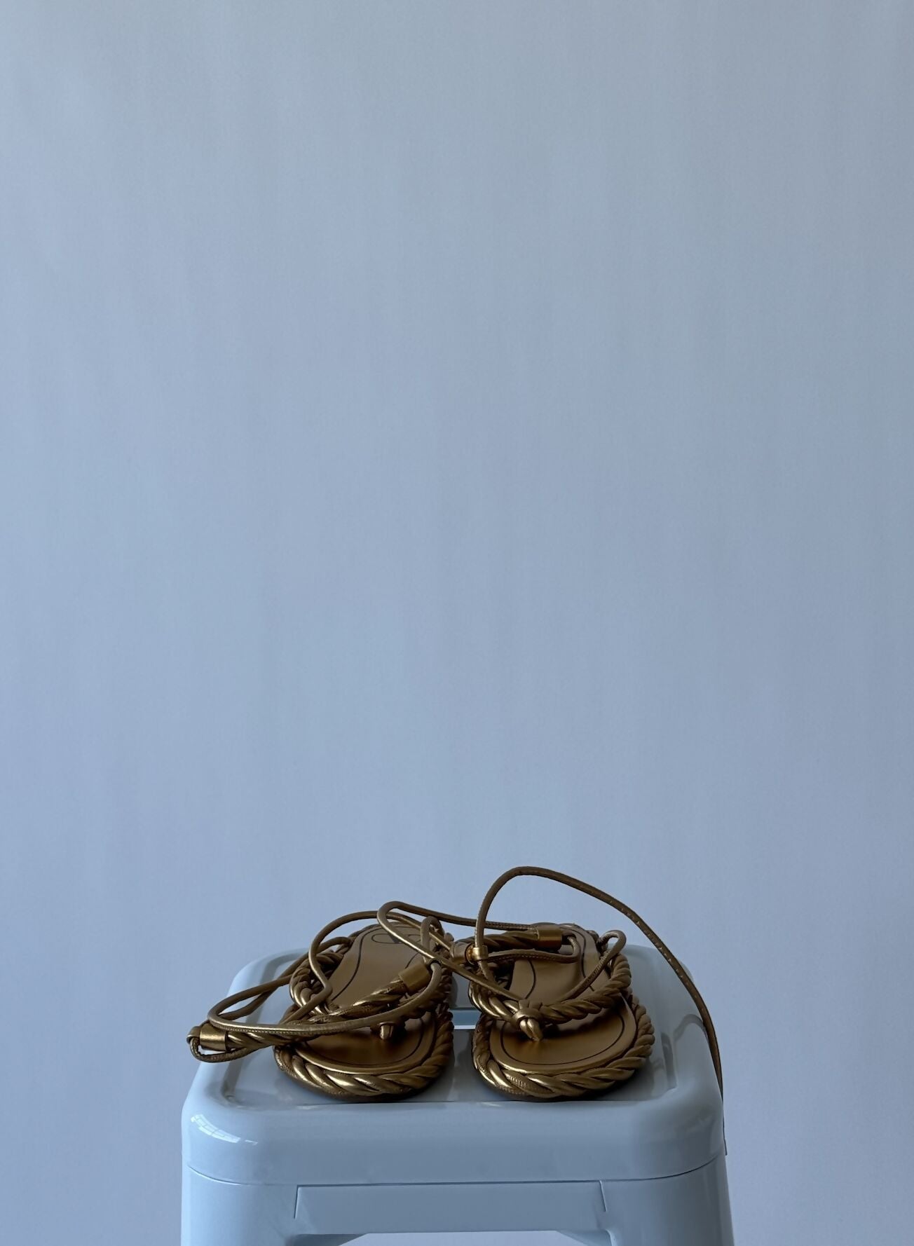 A sold-out pair of EXCHANGE Valentino - THE ROPE SANDALS in size 39, featuring gold rope-like straps, sits neatly on a light gray stool against a plain light background.