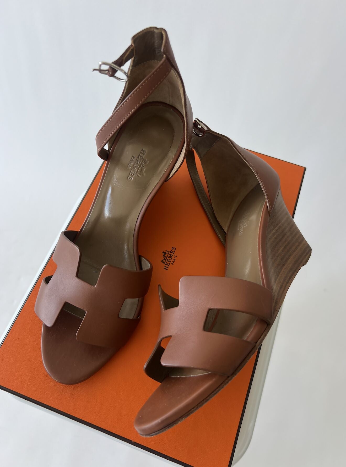 A pair of HERMES Calfskin Legend Wedge Sandals, size EU 39 in brown with cut-out H-shaped straps, displayed on an orange box with dust bags, by EXCHANGE against a white background.