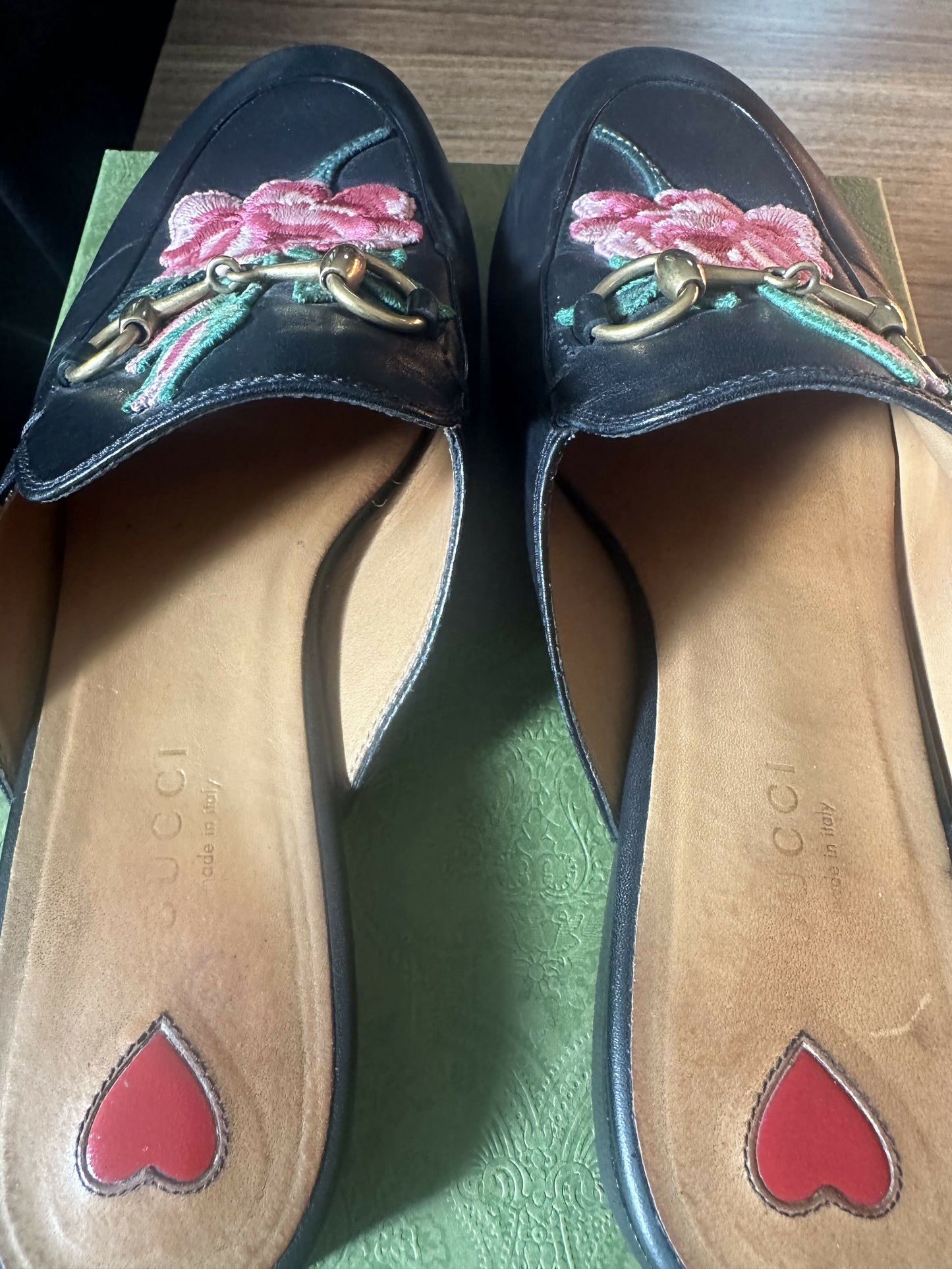 A pair of EXCHANGE Gucci Princetown Black Leather Rose Flower Mules with gold horsebit accents and rose embroidery is displayed on a textured green box; the tan insoles each have a red heart near the heel.