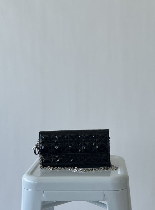 A Lady Dior Pouch by EXCHANGE in black patent leather with silver hardware rests on a white stool against a plain light gray background.