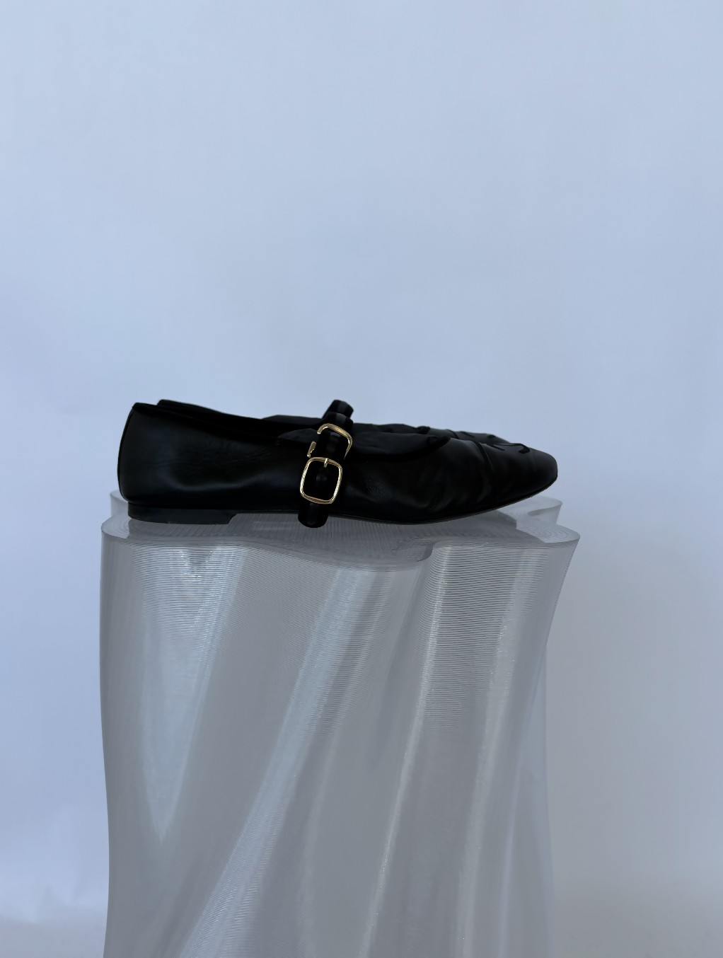 The Chanel Mary Jane flats in Lambskin by EXCHANGE STORE feature soft black leather and a gold buckle, displayed on a translucent, wavy pedestal against a light background—a refined choice for collectors.