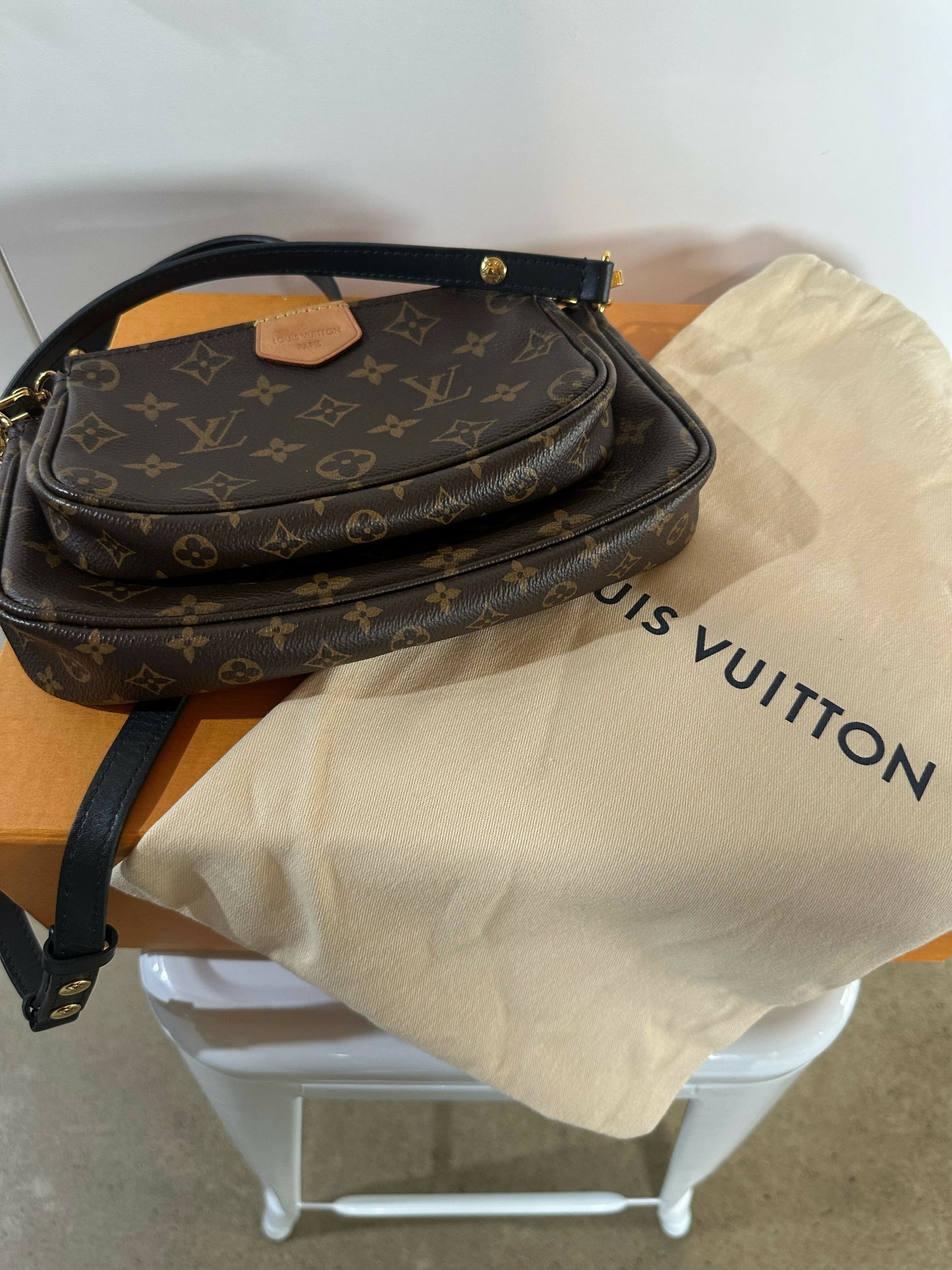 A Louis Vuitton Multi Pochette - Full Set by EXCHANGE is displayed on an orange box with a beige dust bag, all placed on a white chair.