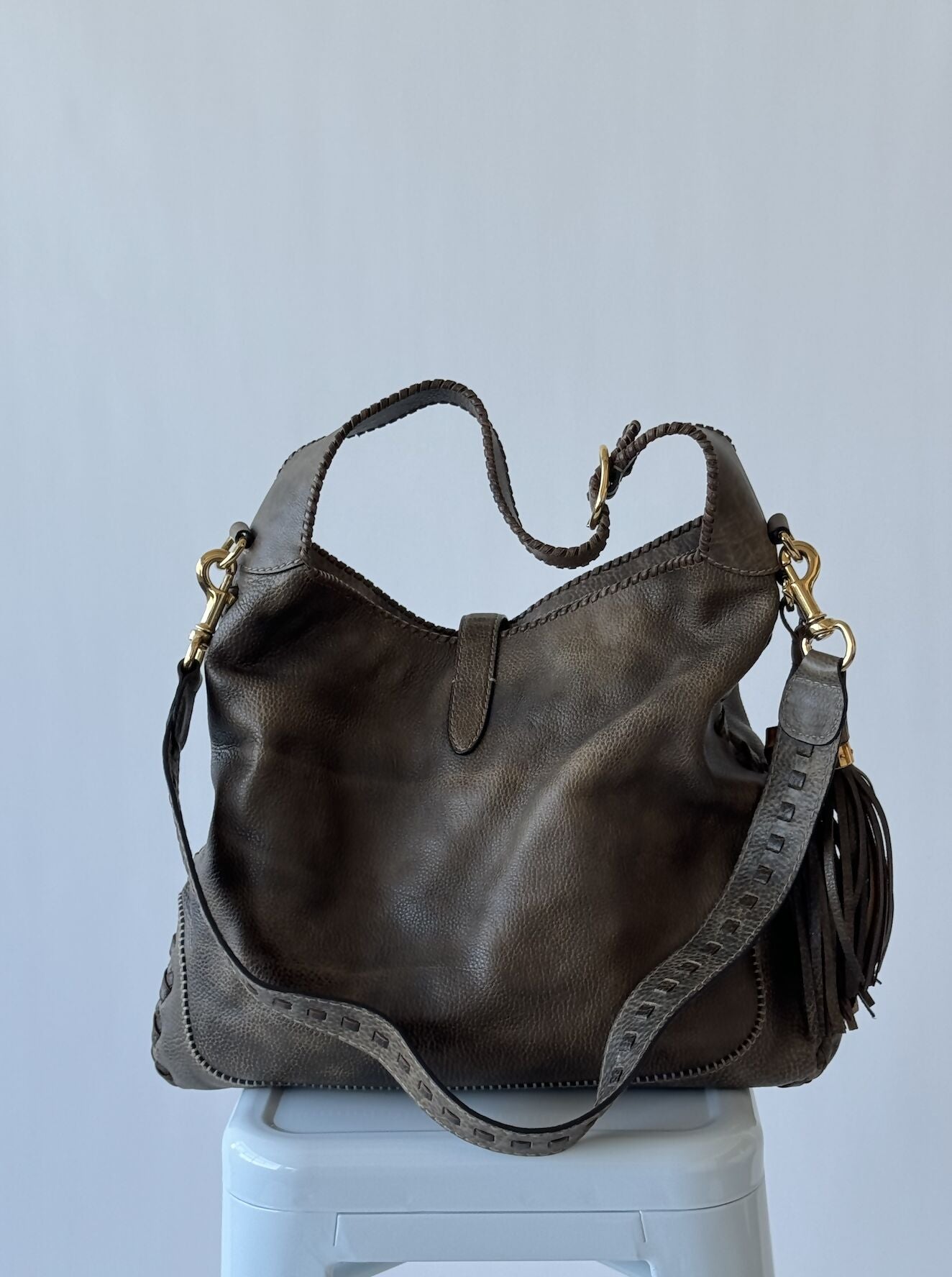 An olive green Gucci Large Jackie Bag by EXCHANGE, featuring a braided strap, gold hardware, and a decorative tassel, is displayed on a white stool against a plain light gray background.