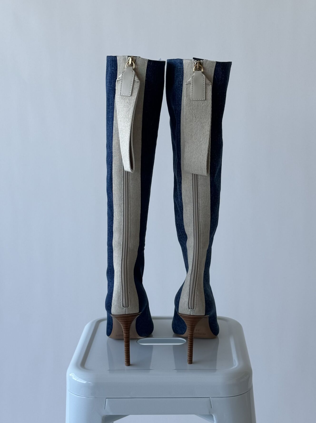 Brand new Jacquemus Les Bottes Cordao knee-high boots in blue and white, with stiletto heels, zippers, and long tags, are displayed upright on a white stool against a light gray background. Brand Name: EXCHANGE.