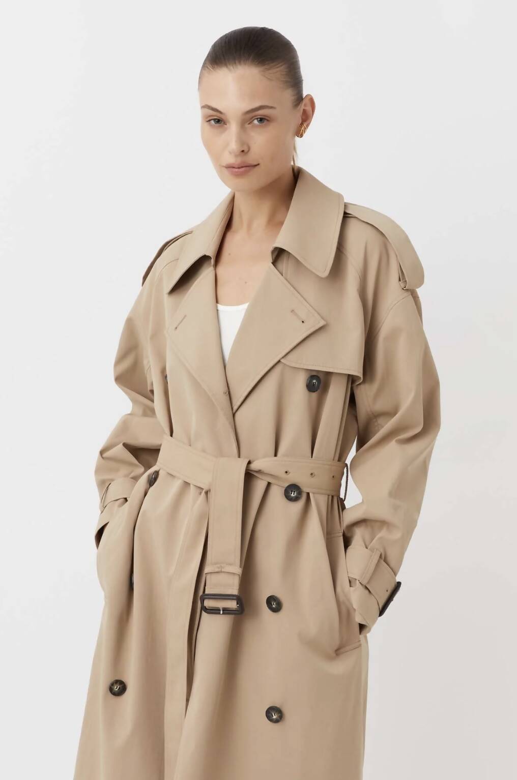A woman wears an extra small EXCHANGE Evans Trench Coat by Camilla & Marc in light beige, with a belt and black buttons. The coat, worn once and in excellent condition, stands out against a plain white background.