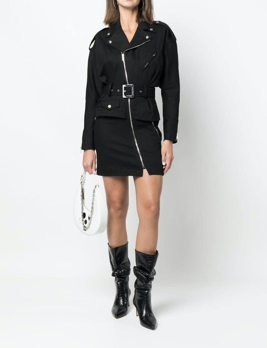 A woman wearing the Alexandre Vauthier Fastened biker mini dress in 100% leather by EXCHANGE, paired with black pointed-toe boots and a white chain-strap handbag, stands against a plain white background.