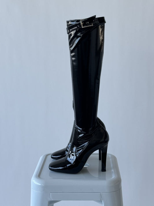 A pair of SAINT LAURENT Patent Stretch Crystal Buckle Lala boots by EXCHANGE are displayed upright on a white stool against a plain light background.