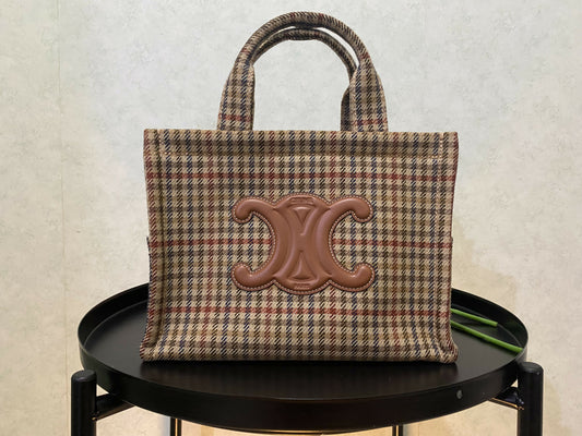 An Authentic Celine handbag by EXCHANGE with plaid design and double handles, featuring a prominent brown leather emblem, rests in excellent condition on a round black table against a light textured background.