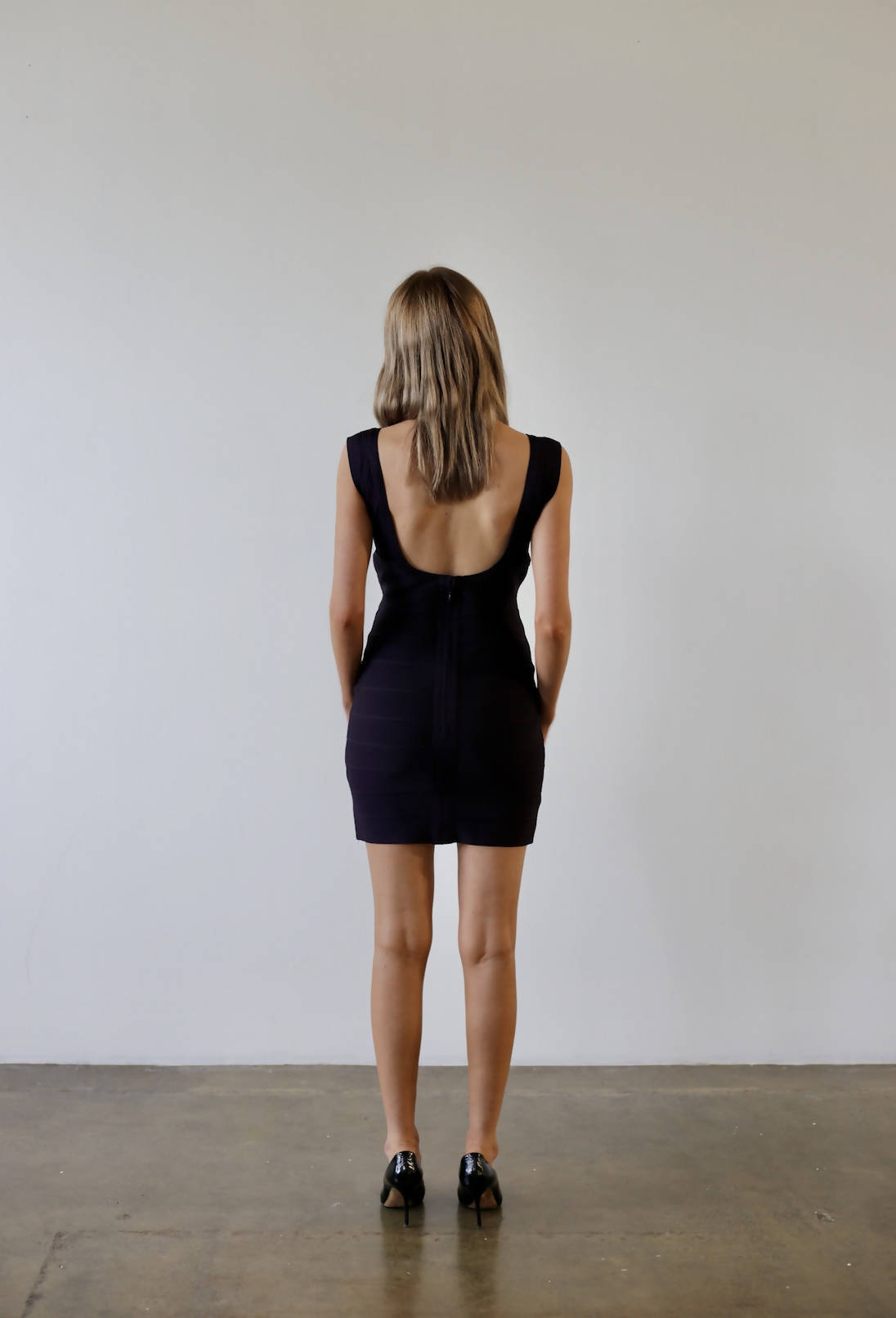 A person with long hair, back facing the camera, wears a sleeveless black dress similar to an EXCHANGE Herve Leger dress and sleek black shoes. They stand confidently on a concrete floor against a plain, light-colored wall.