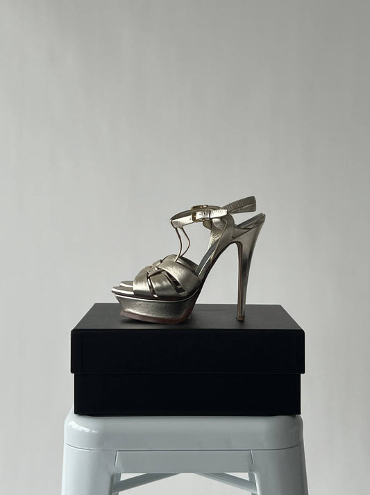 A Saint Laurant Tribute Heel 105 by EXCHANGE, size 38, metallic with ankle straps and a 105mm heel, is shown on a black box placed on a white stool against a plain light gray background.