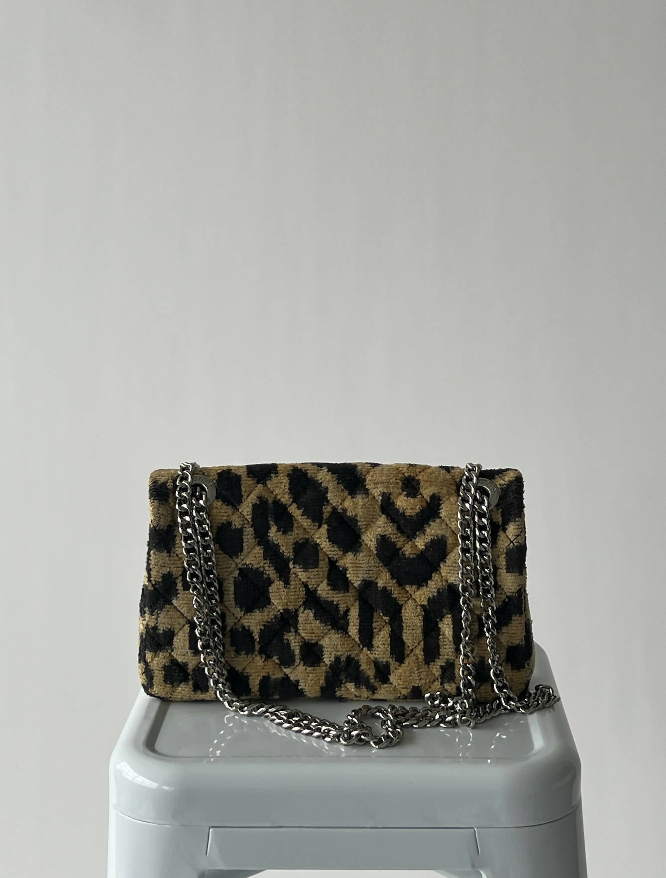 A used EXCHANGE Rough Studios Bag with a gold and black geometric animal print, silver chain strap, and dustbag is displayed on a white stool against a plain light gray background.