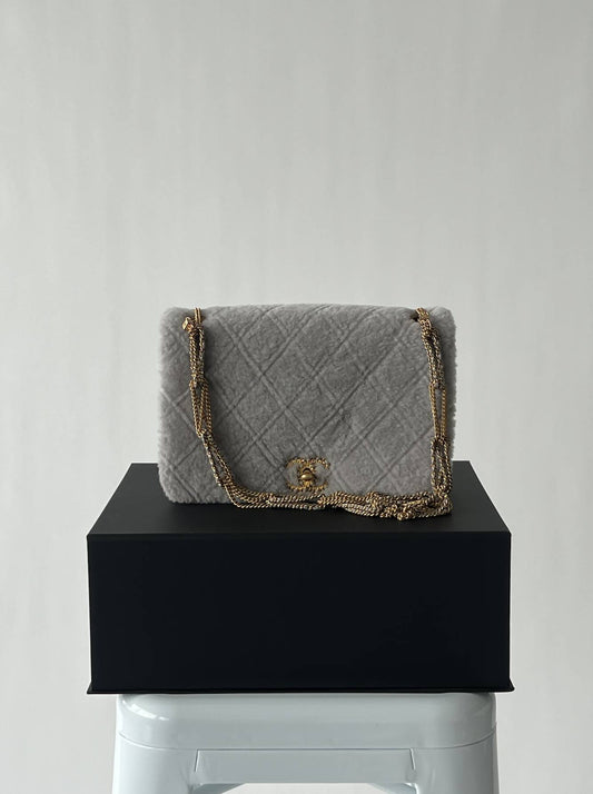 Chanel’s 2022 Métiers d’Art collection - Flap Bag by EXCHANGE: a light gray shirling and suede design with gold chain strap and logo, displayed on a black box on a white stool—a collector’s dream.