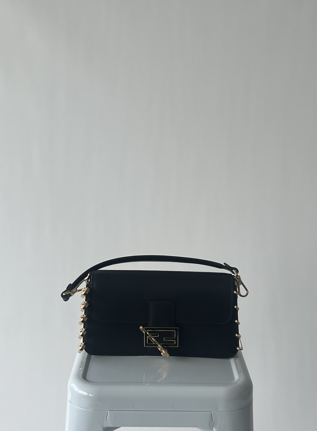 The EXCHANGE FENDI X Versace Fendace Baguette Shoulder Bag, featuring black leather, gold hardware, a top handle, and a front clasp, sits on a white stool against a light gray background—encapsulating Italian fashion.