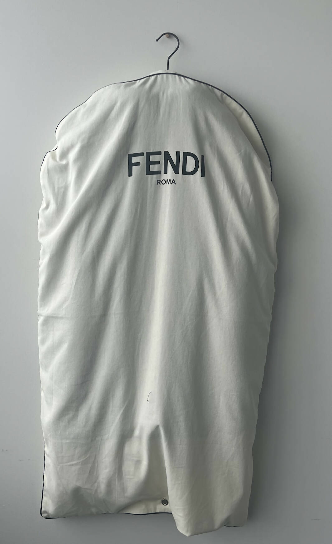 A white garment bag with a black EXCHANGE logo hangs on a metal hanger against a light gray wall, hinting at the luxury of an EXCHANGE Fendi Jacket Black shearling jacket inside.