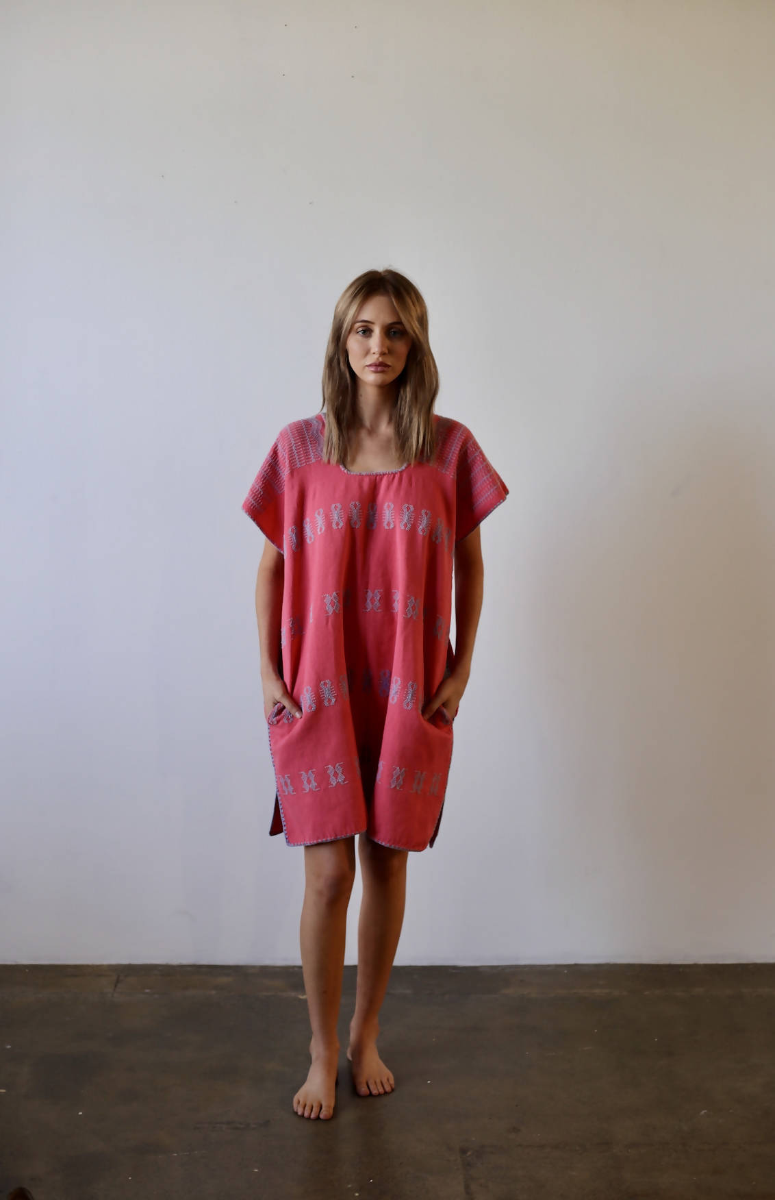A person stands barefoot on a concrete floor against a plain white wall, wearing the unworn Pippa Holt Kaftan by EXCHANGE. This one-size-fits-all kaftan is loose-fitting with short sleeves and pockets, featuring white patterns for a casual and relaxed look.