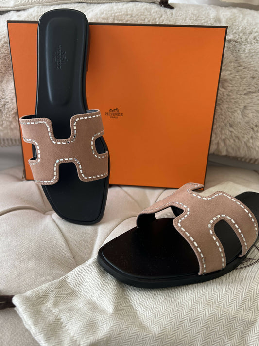 A pair of Lulu Hermes Oran sandals NEW “SALE” $1500, with tan suede straps and white stitching, are displayed on a beige surface—one on an orange Hermès box, the other on a fabric pouch.