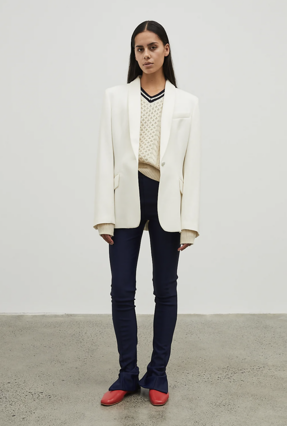 A person with long dark hair stands indoors against a white wall, wearing the EXCHANGE Maggie Marylin 01 Legging (size small, gently worn), styled with a white blazer, cream sweater, and red shoes for a minimal modern look.