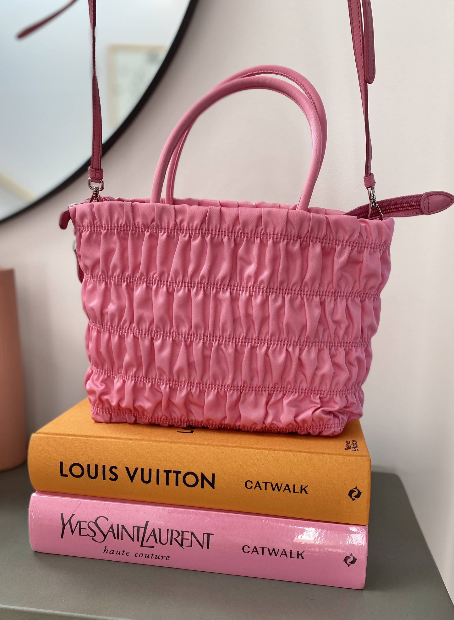 The Prada Pink Nylon Tote by EXCHANGE, featuring handles, a nappa leather shoulder strap, and an enameled metal triangle logo, rests atop two fashion books—Louis Vuitton Catwalk (orange) and Yves Saint Laurent Catwalk (pink)—by a mirror.