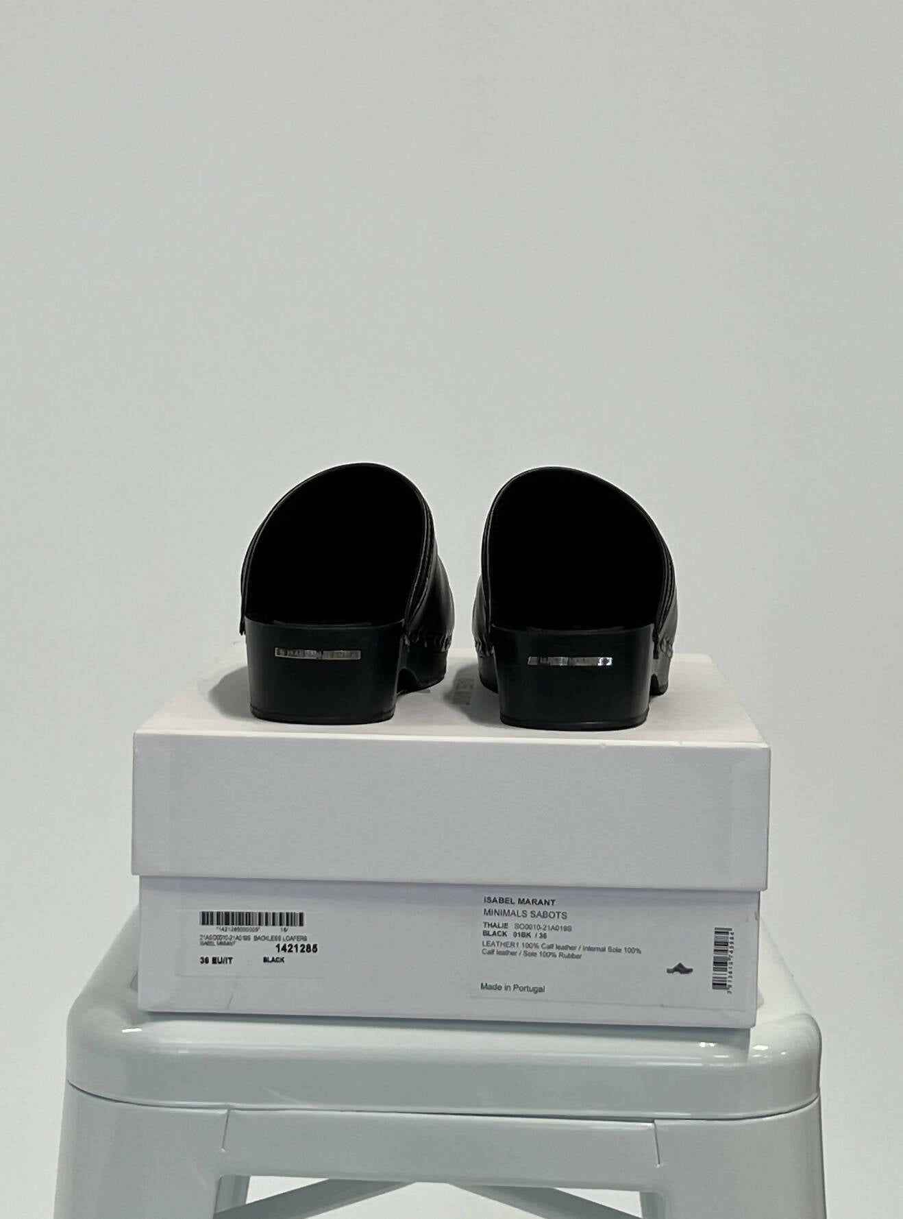The EXCHANGE Isabel Marant Minimals Sabots, black slip-on mules, are shown with heels forward on their original box atop a light gray stool against a plain white background.