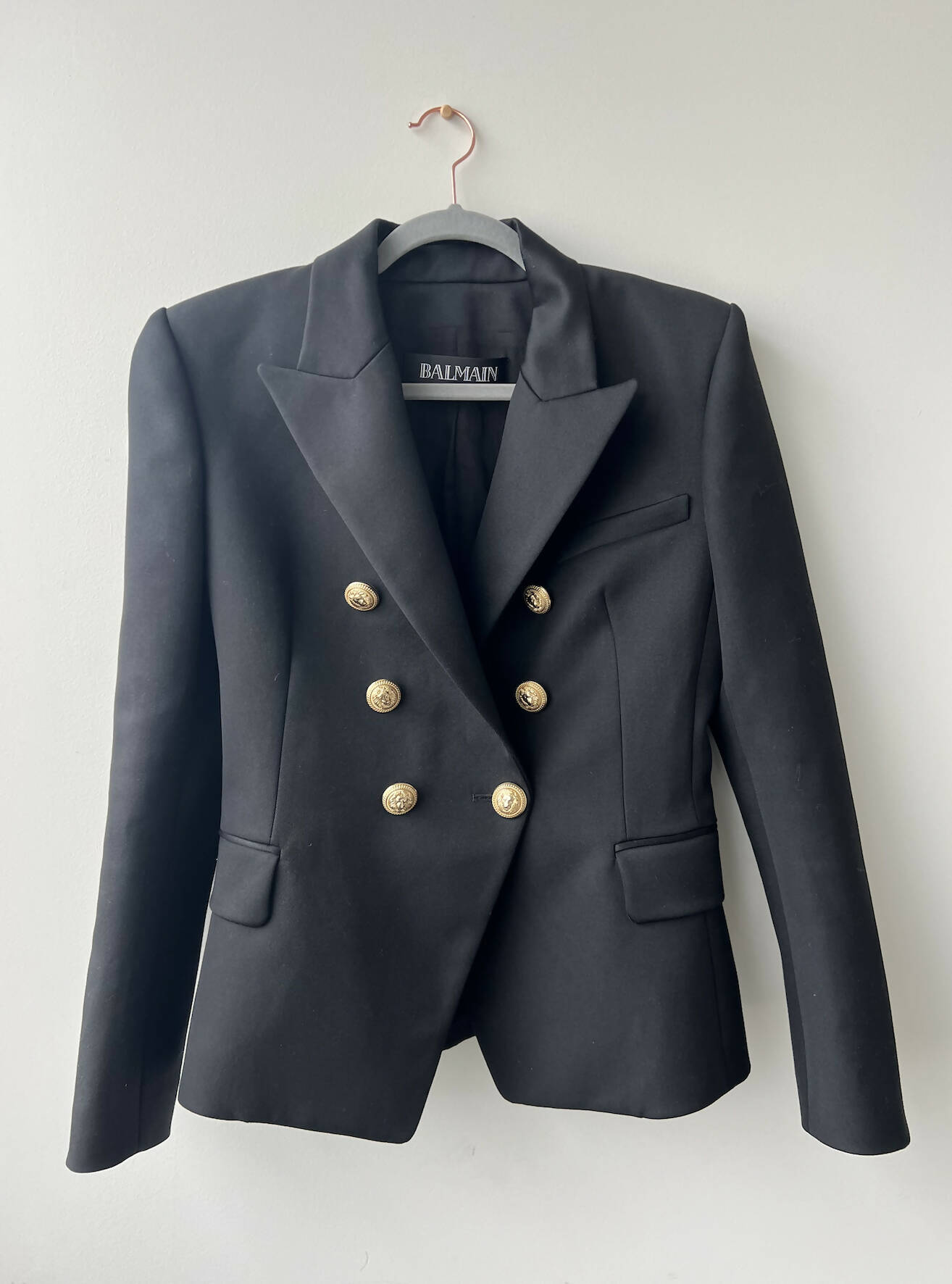 The EXCHANGE Balmain Blazer Black features gold buttons, structured shoulders, and peaked lapels. Made in Slovakia, it hangs on a pink hanger against a plain light-colored wall.