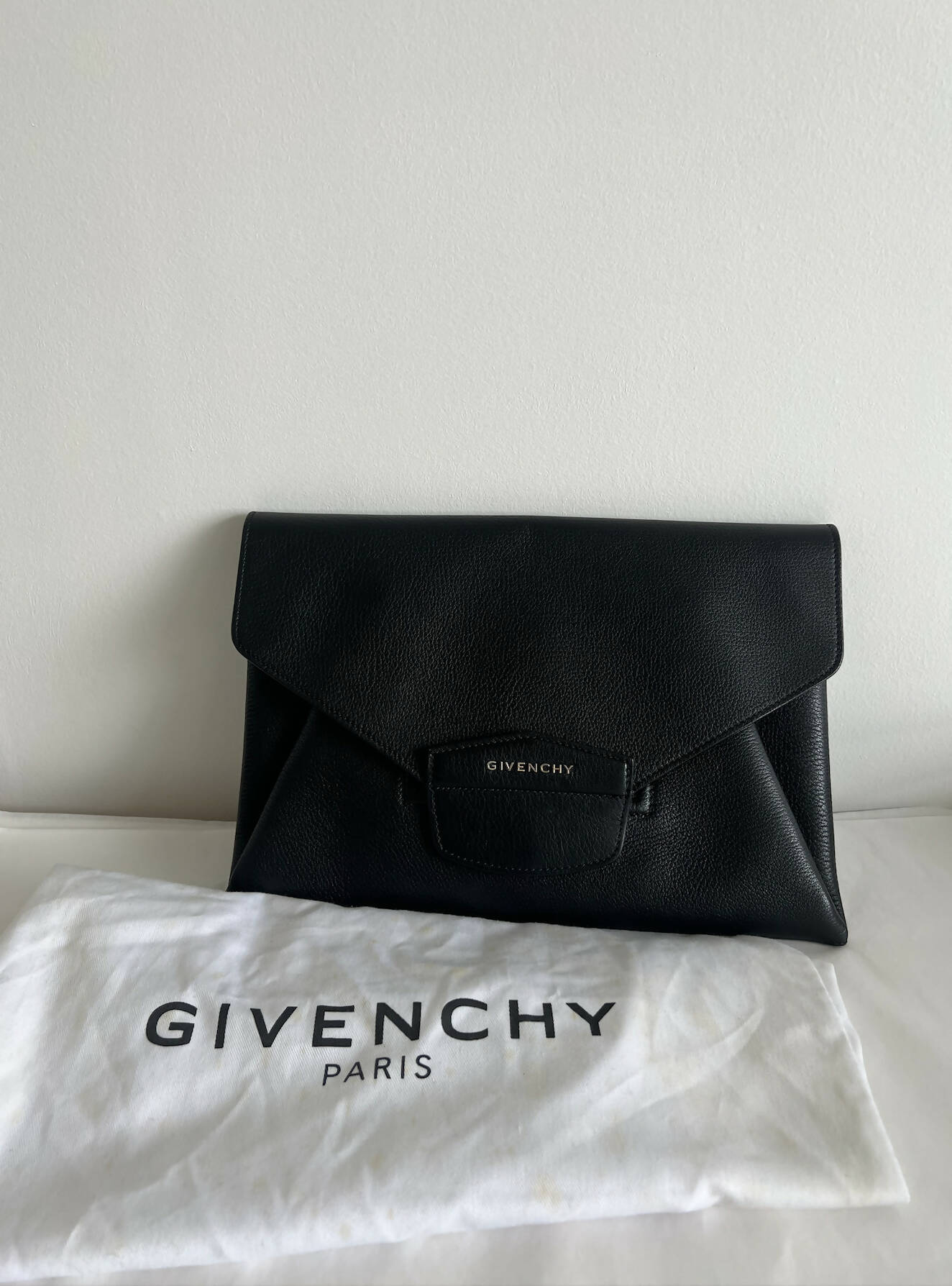 A pre-owned GIVENCHY ANTIGONA ENVELOPE CLUTCH BAG by EXCHANGE in black goatskin leather is displayed upright against a white wall, with its white Givenchy Paris dust bag laid flat in front.