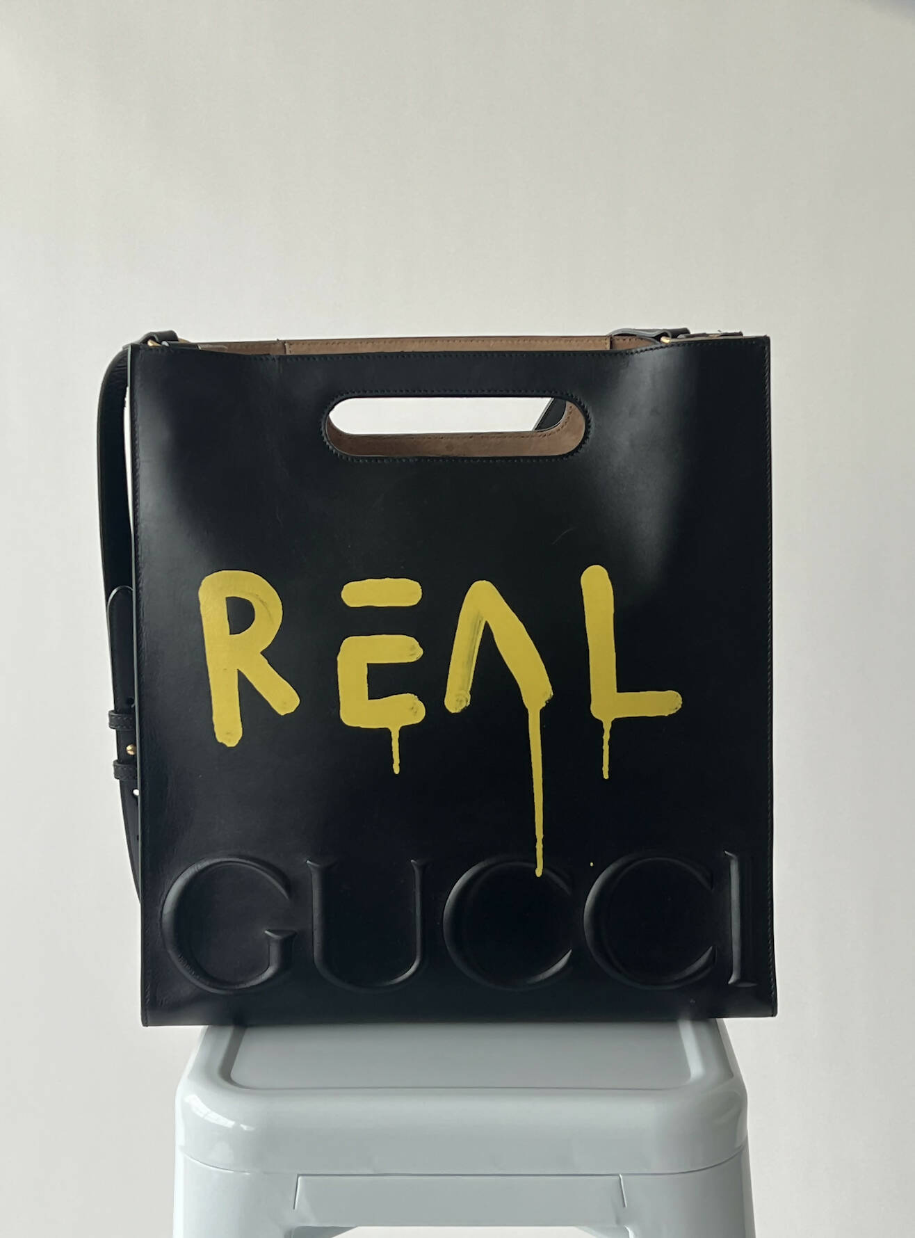 A black calfskin GUCCI Black GucciGhost Print Medium XL Tote Bag by EXCHANGE sits on a stool, featuring yellow graffiti "REAL" with a dripping A and embossed GUCCI at the bottom, plus cut-out handles.