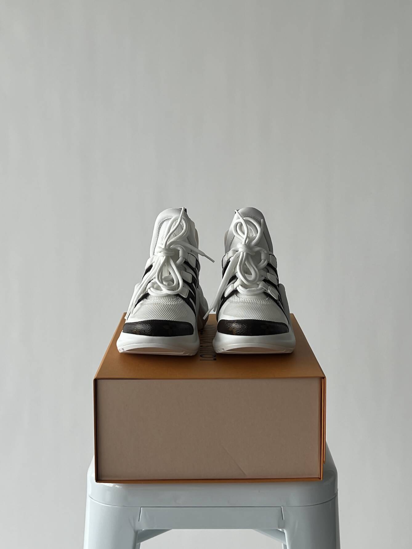 A pair of EXCHANGE LV Archlight Sneakers in white and black, size 38 with white laces, are set side by side on a closed brown shoe box atop a white stool against a plain light gray background. They are in fantastic condition.