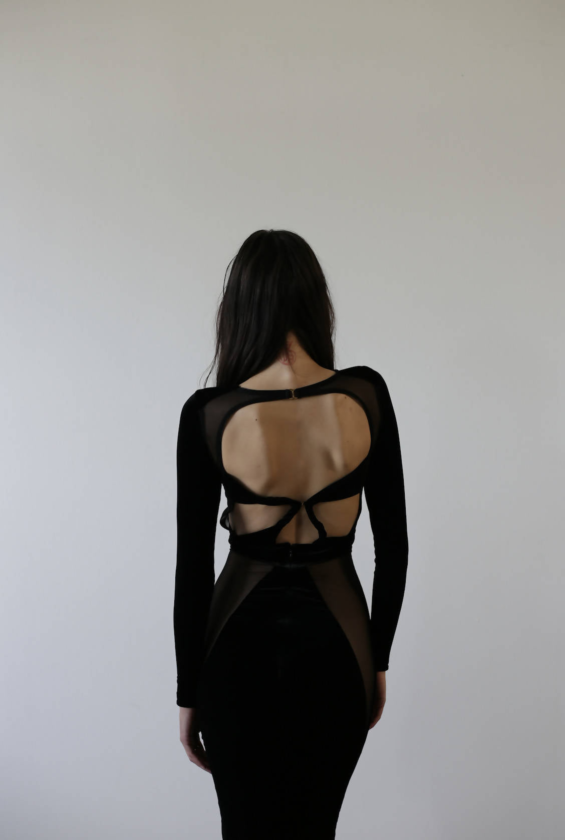 A person with long dark hair faces away from the camera in a stunning EXCHANGE Bao Trenchi Dress, featuring an open back and elegant crisscrossing straps, set against a plain, light-colored wall.