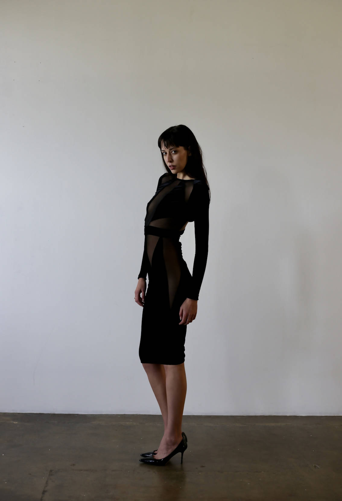 A woman wearing an EXCHANGE Bao Trenchi Dress, featuring sheer panels and a fitted mesh design, poses against a plain white background. She stands sideways, slightly angled toward the camera. Her long hair and black heels complete the minimal studio-like setting.
