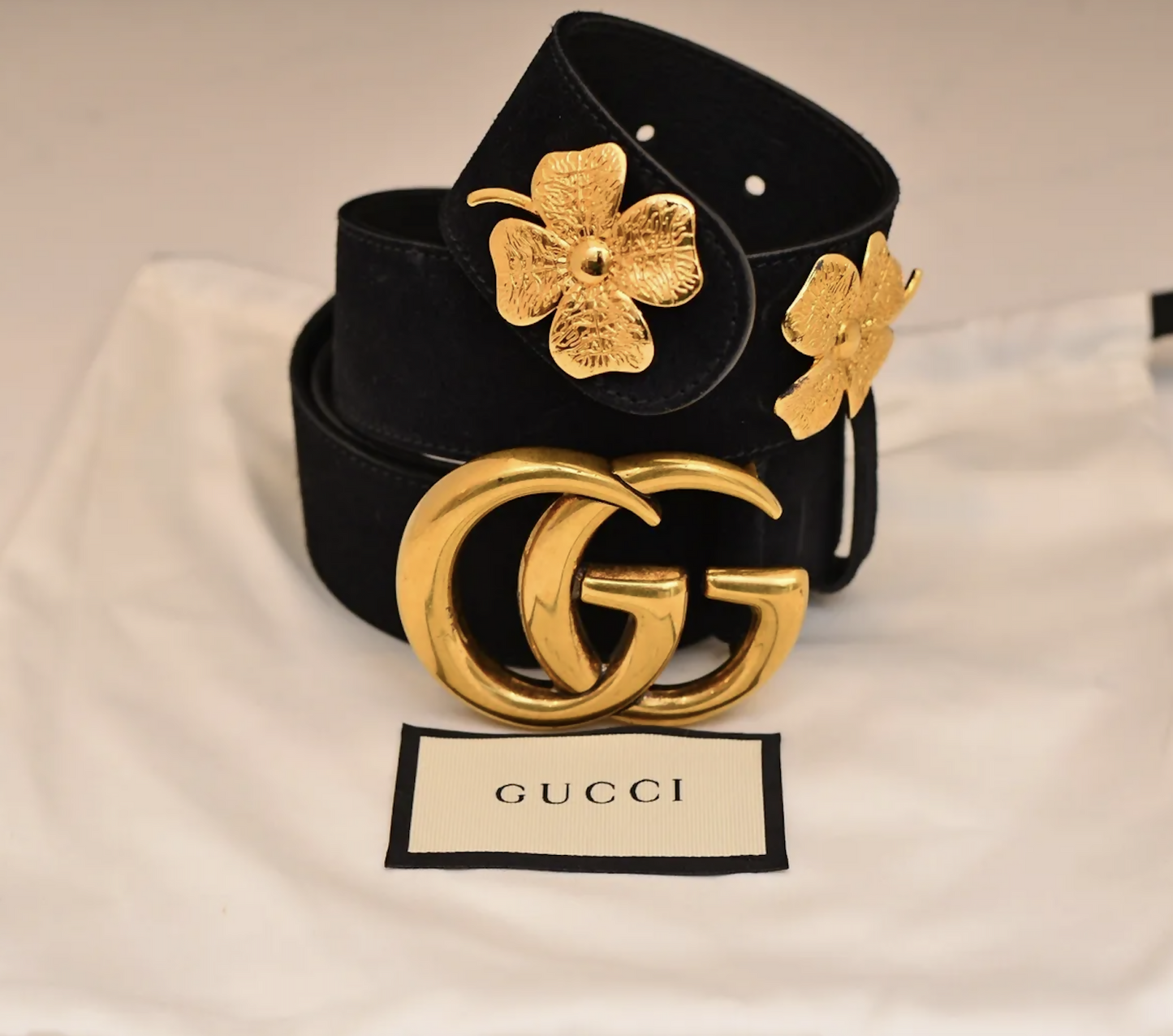 The EXCHANGE Gucci Clover ‘Flore Blooms’ Belt features a black strap with a bold gold double G buckle and gold floral accents, shown on a white Gucci dust bag with the brand label visible.