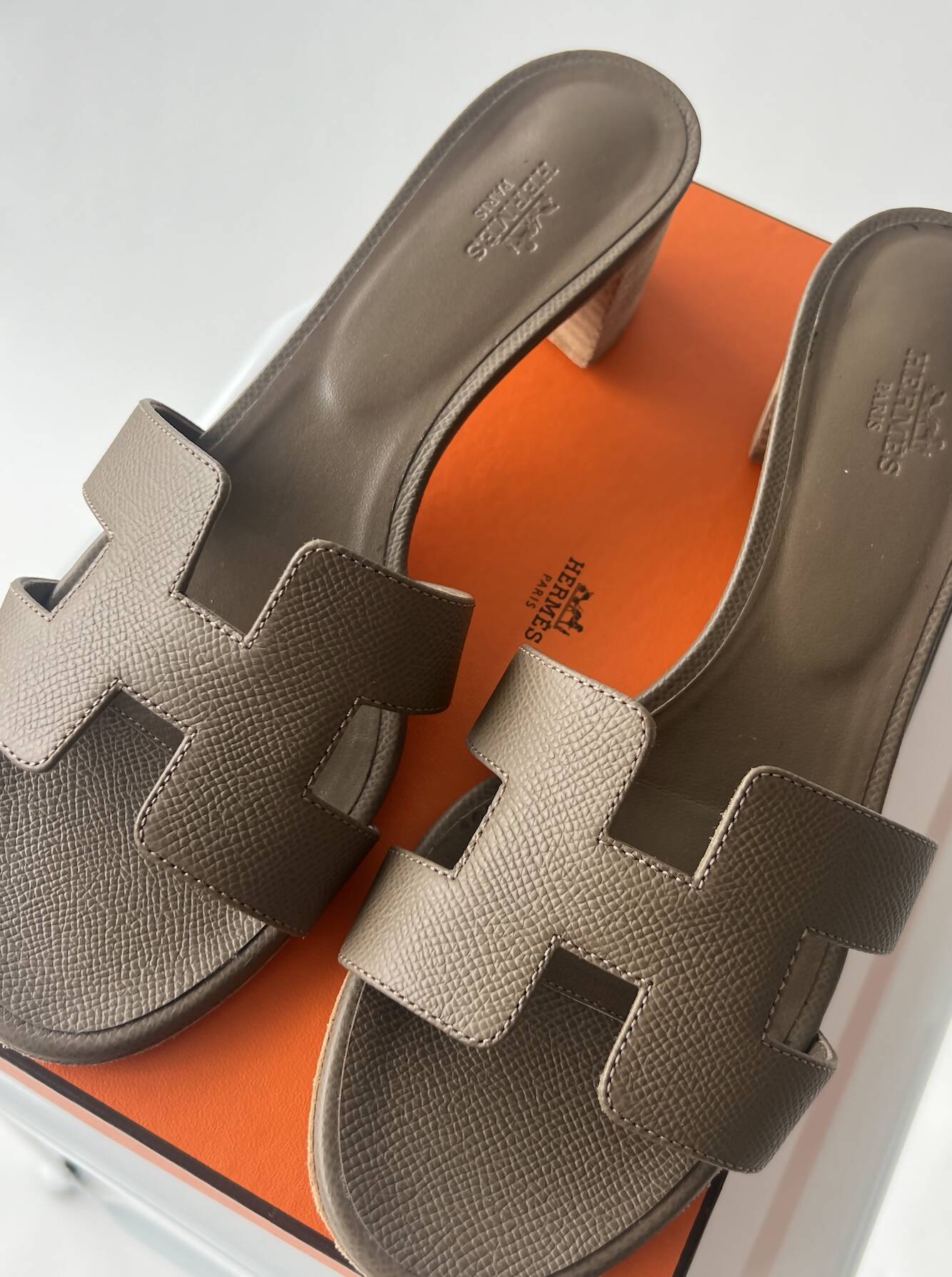 A pair of Hermès Healed Oasis Sandals - Full Set by EXCHANGE, size 39.5 in taupe textured leather with H cutout design, displayed atop an orange Hermès box on a white background.