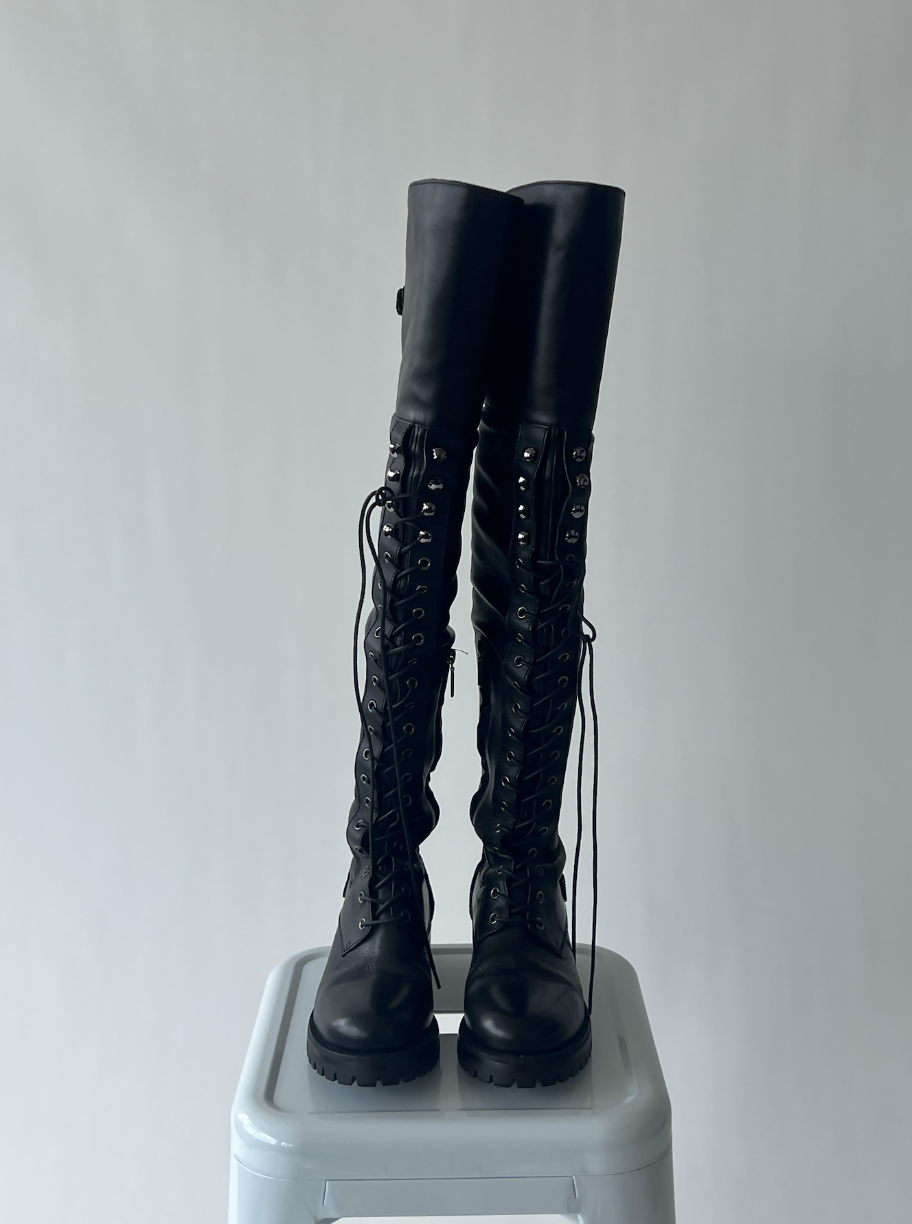 A pair of EXCHANGE Dolce & Gabbana Thigh High Combat Boots in size 38.5, black with chunky soles and lace-up design, placed side by side on a light gray stool against a plain light background.
