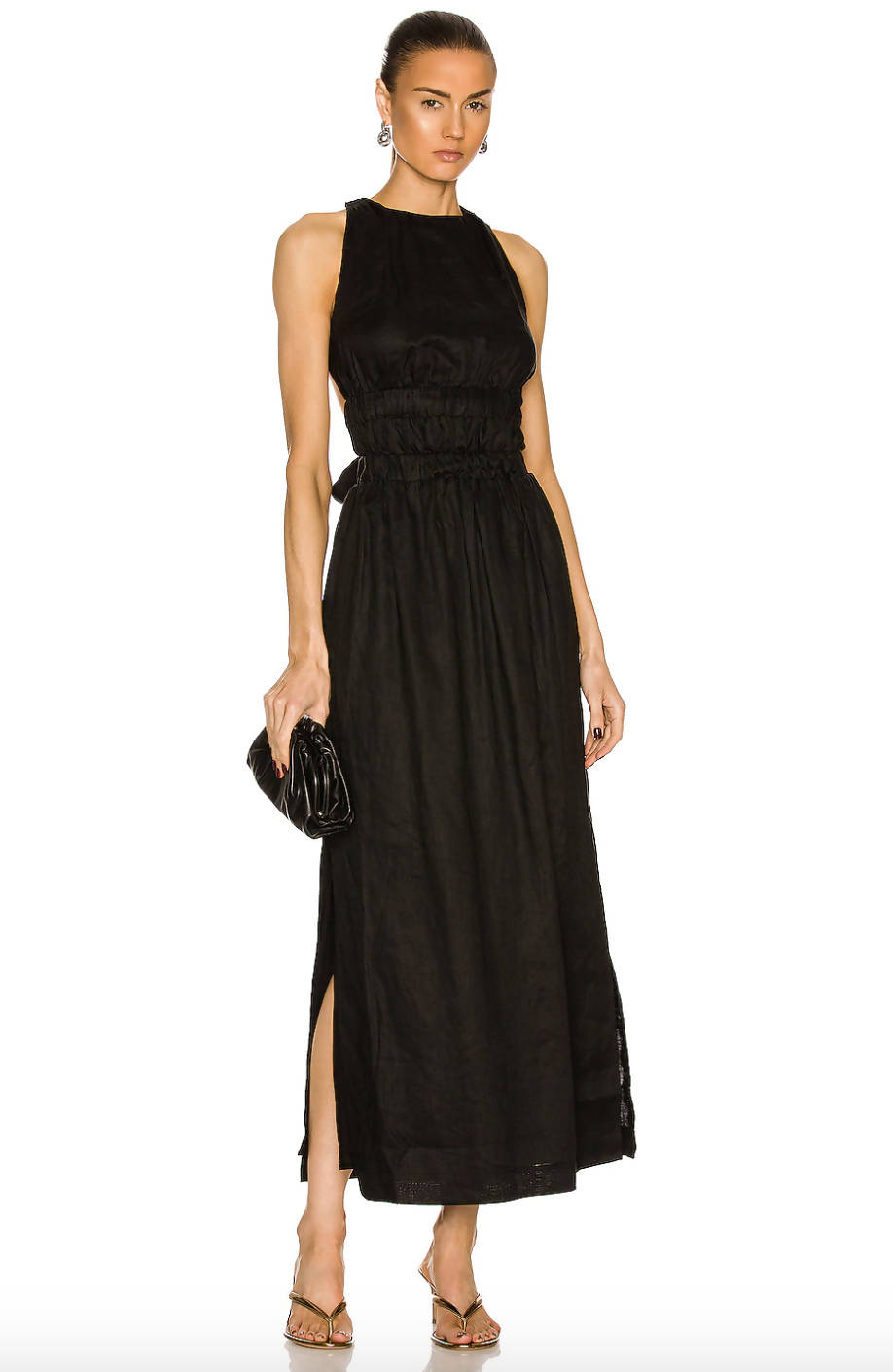 A woman in the EXCHANGE SIR. Vilma Cross Back Gown—a sleeveless, ankle-length black linen dress with elastic waist and side slit—holds a black clutch, wears heel sandals, has her hair tied back, and accessorizes with earrings.