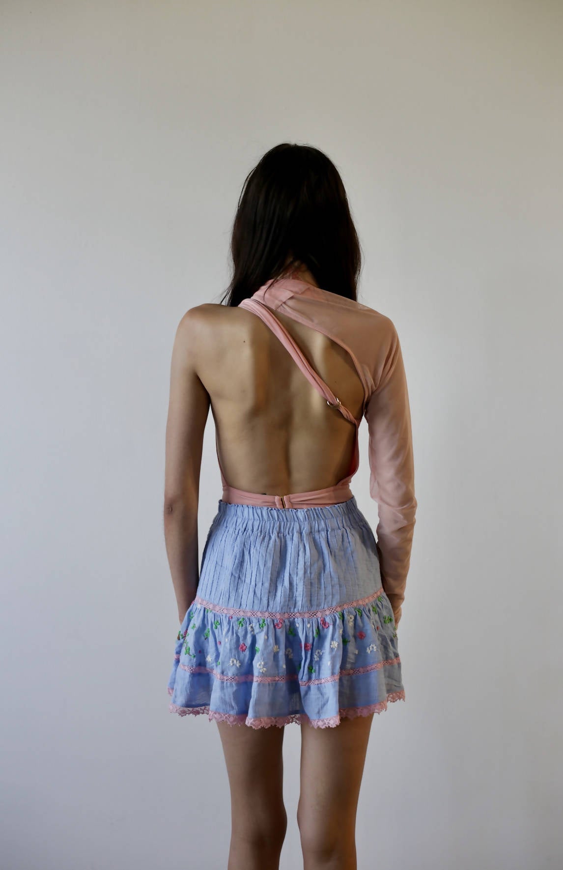 A person with long dark hair, facing away, wears an unworn EXCHANGE Bao Tranchi Body Suit in size 36—pink, asymmetrical with one sleeve and open back—paired with a light blue ruffled skirt with colorful trim against a plain background.