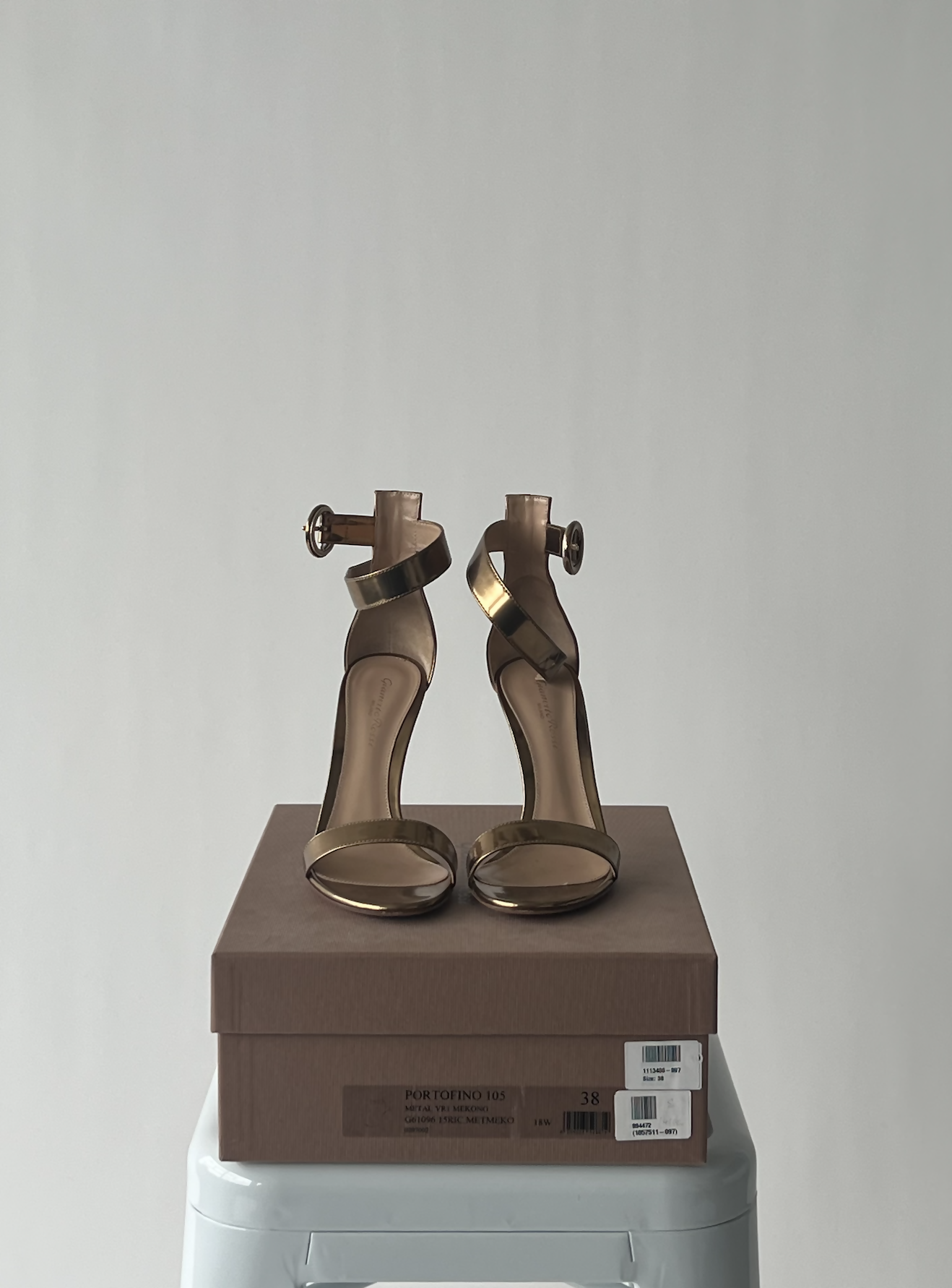 A pair of Gianvito Rossi Gold Leather Portofino Ankle Strap Sandals by EXCHANGE rests on a brown shoebox placed atop a white stool, set against a plain light gray background.
