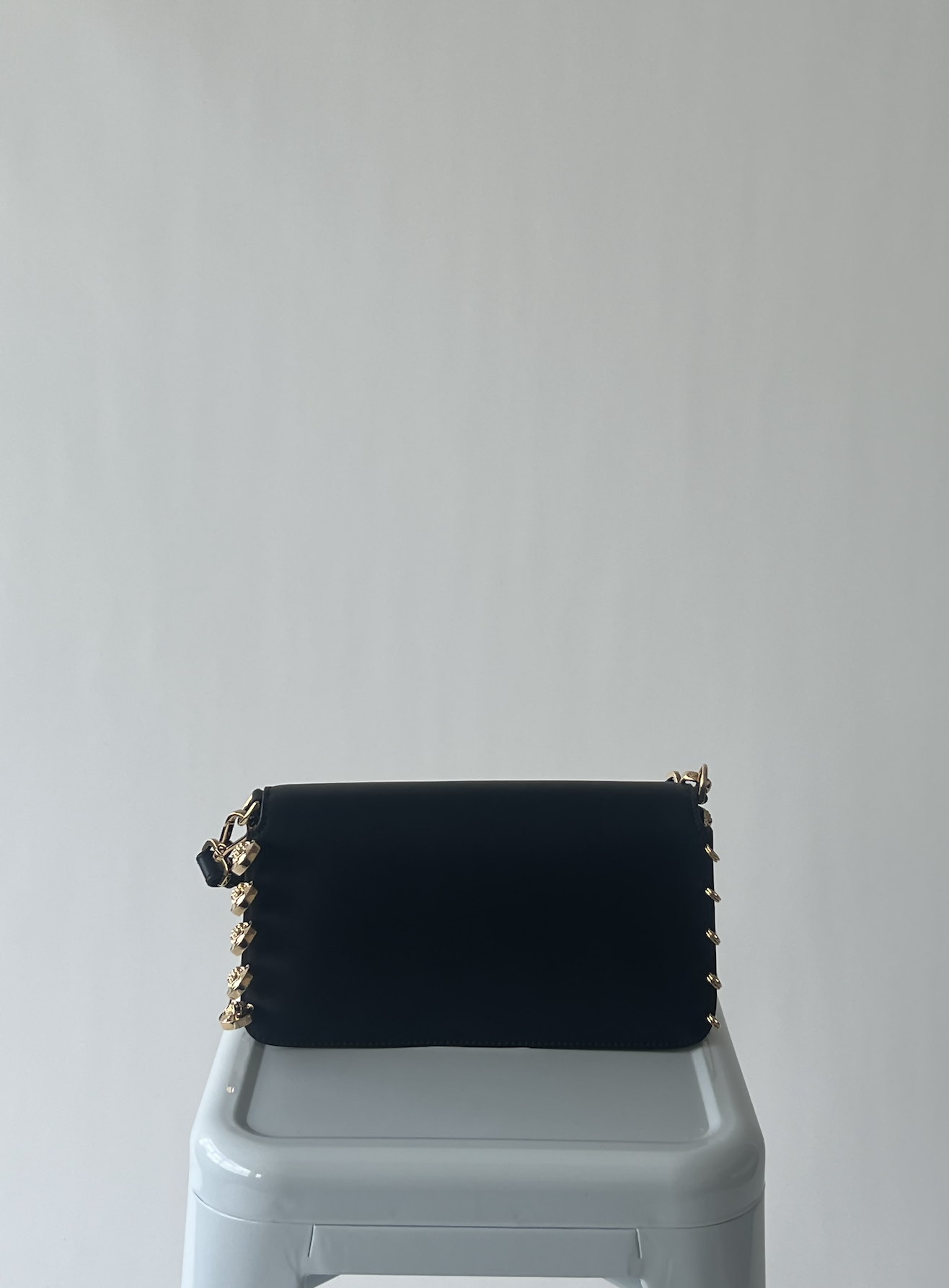 The FENDI X Versace Fendace Baguette Shoulder Bag by EXCHANGE, a black rectangular luxury piece with gold studs on the sides, rests on a white stool against a plain light gray background.