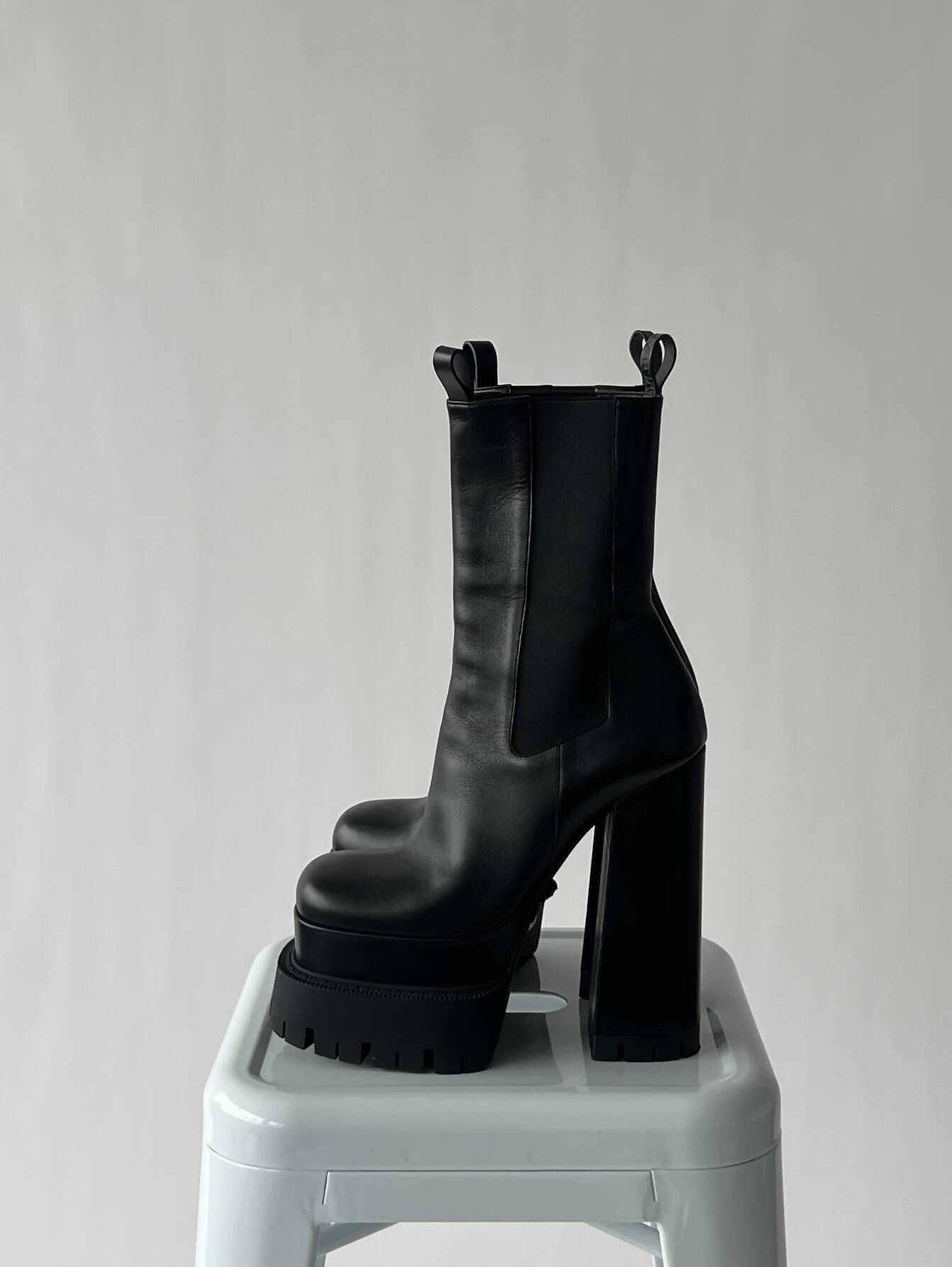 A pair of EXCHANGE VERSACE Aevitas Platform Boots - FULL SET, size 39, with chunky high heels and pull tabs, is displayed on a white stool against a plain light background.