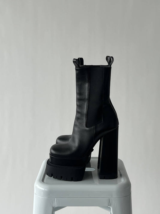 A pair of EXCHANGE VERSACE Aevitas Platform Boots - FULL SET, size 39, with chunky high heels and pull tabs, is displayed on a white stool against a plain light background.
