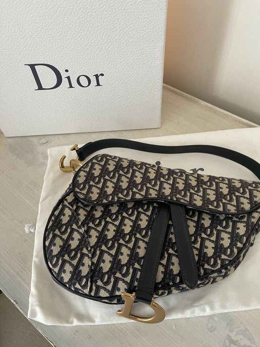 The EXCHANGE Christian Dior Saddle Oblique Medium Shoulder Bag in Jacquard, featuring a black and beige monogram pattern, rests on a white dust bag beside a textured white Dior box atop light wood—a collector's essential.