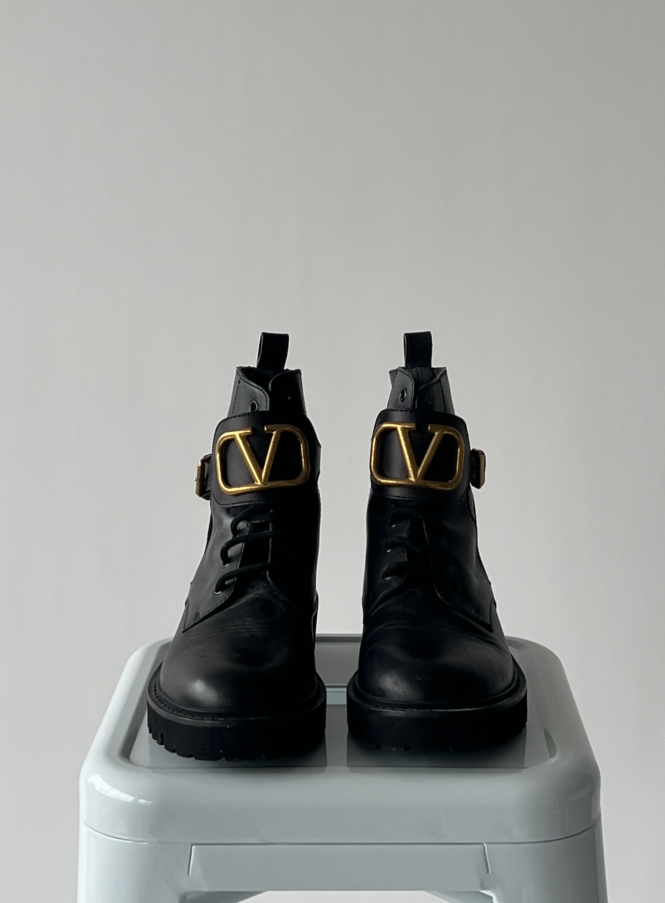 A pair of EXCHANGE Valentino Garavani VLOGO leather ankle boots with chunky soles and gold V-shaped buckles, displayed on a light gray stool against a plain, light background.