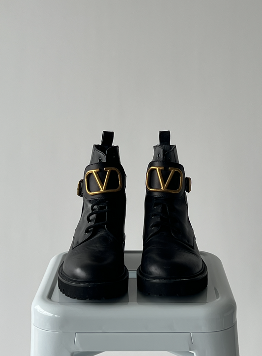 A pair of EXCHANGE Valentino Garavani VLOGO leather ankle boots with chunky soles and gold V-shaped buckles, displayed on a light gray stool against a plain, light background.