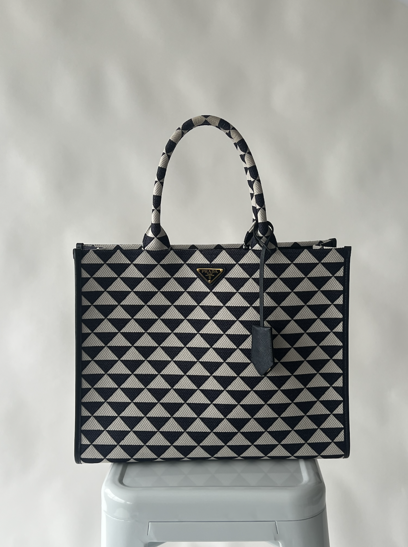 A Large Prada Symbole embroidered fabric handbag by EXCHANGE, featuring top handles and a small triangular logo, is displayed on a white stool against a light, blurred background.