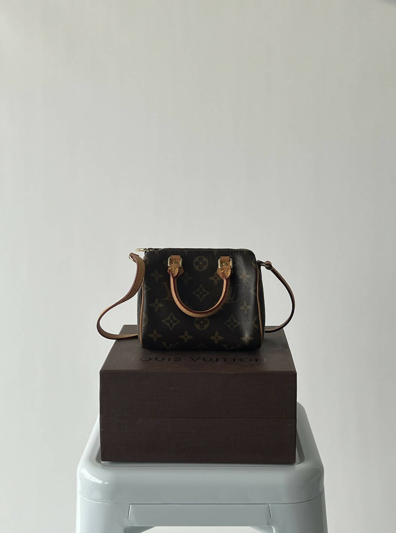 A Louis Vuitton Speedy Nano by EXCHANGE, featuring tan handles and a shoulder strap, rests on a brown box placed on a white stool against a plain light background.