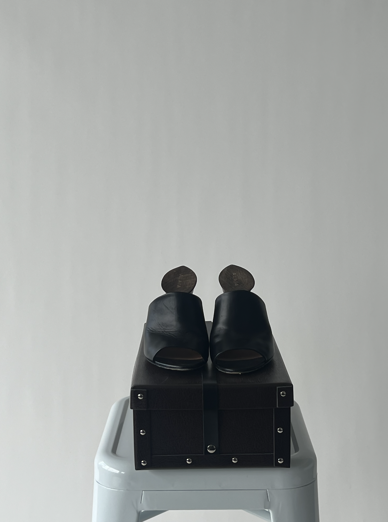 The Alaïa Cut Out Sculpture Mules - Noir by EXCHANGE, featuring open toes and shoehorns, rest on a black box atop a white stool against a plain gray background, exuding French couture elegance.