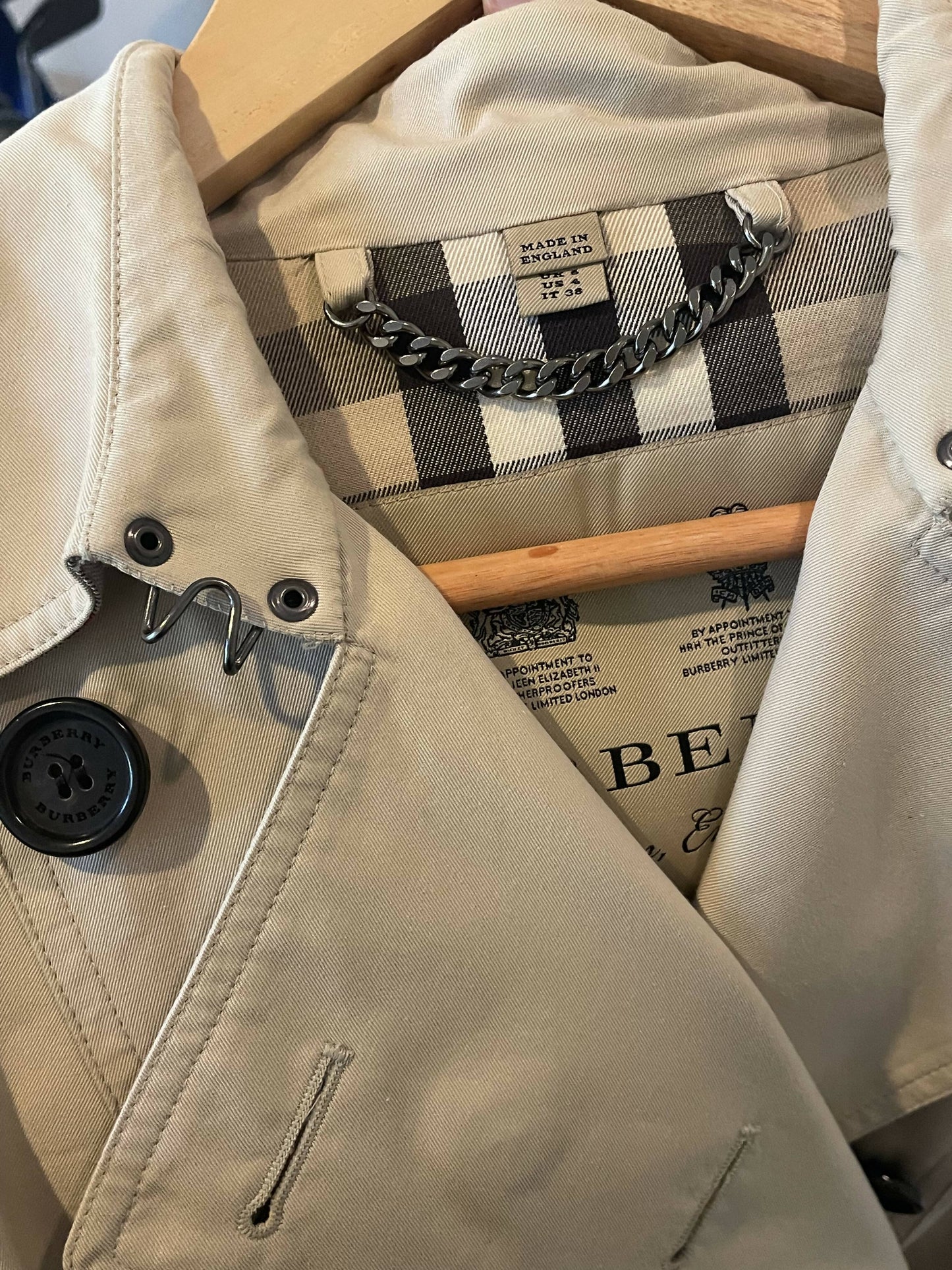 A close-up of an EXCHANGE Burberry - Mid-length Chelsea Heritage Trench Coat UK Size 6 in beige on a hanger, revealing the vintage check lining, hanging chain, and "Made in England" and "Burberry" labels.