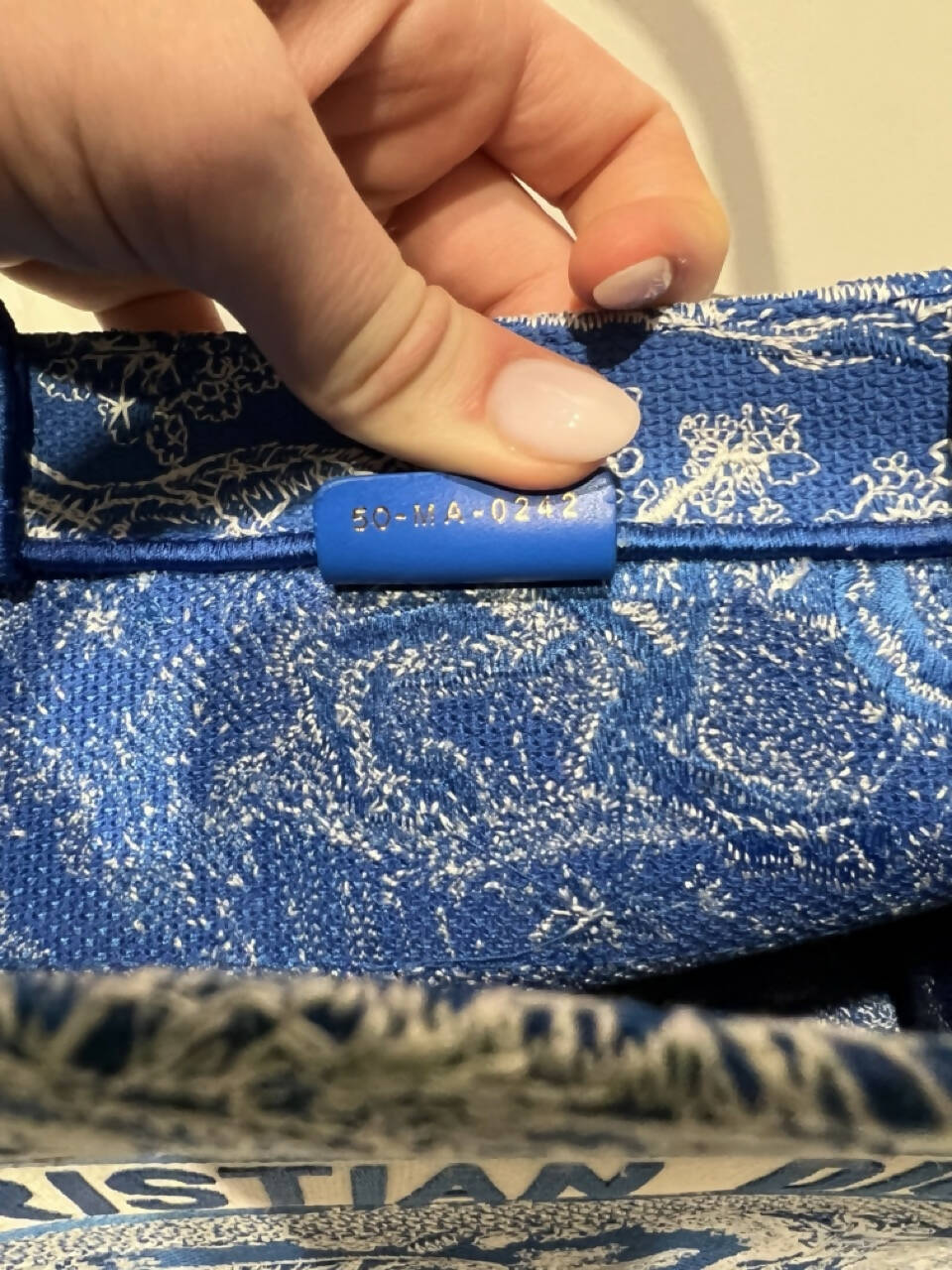 A hand with light pink nails holds open a Dior Book Tote Medium Dioriviera Toile De Jouy Fluorescent Blue by Jess Perham, showing a blue leather tag inside stamped with the code 50-MA-0242 in gold letters.